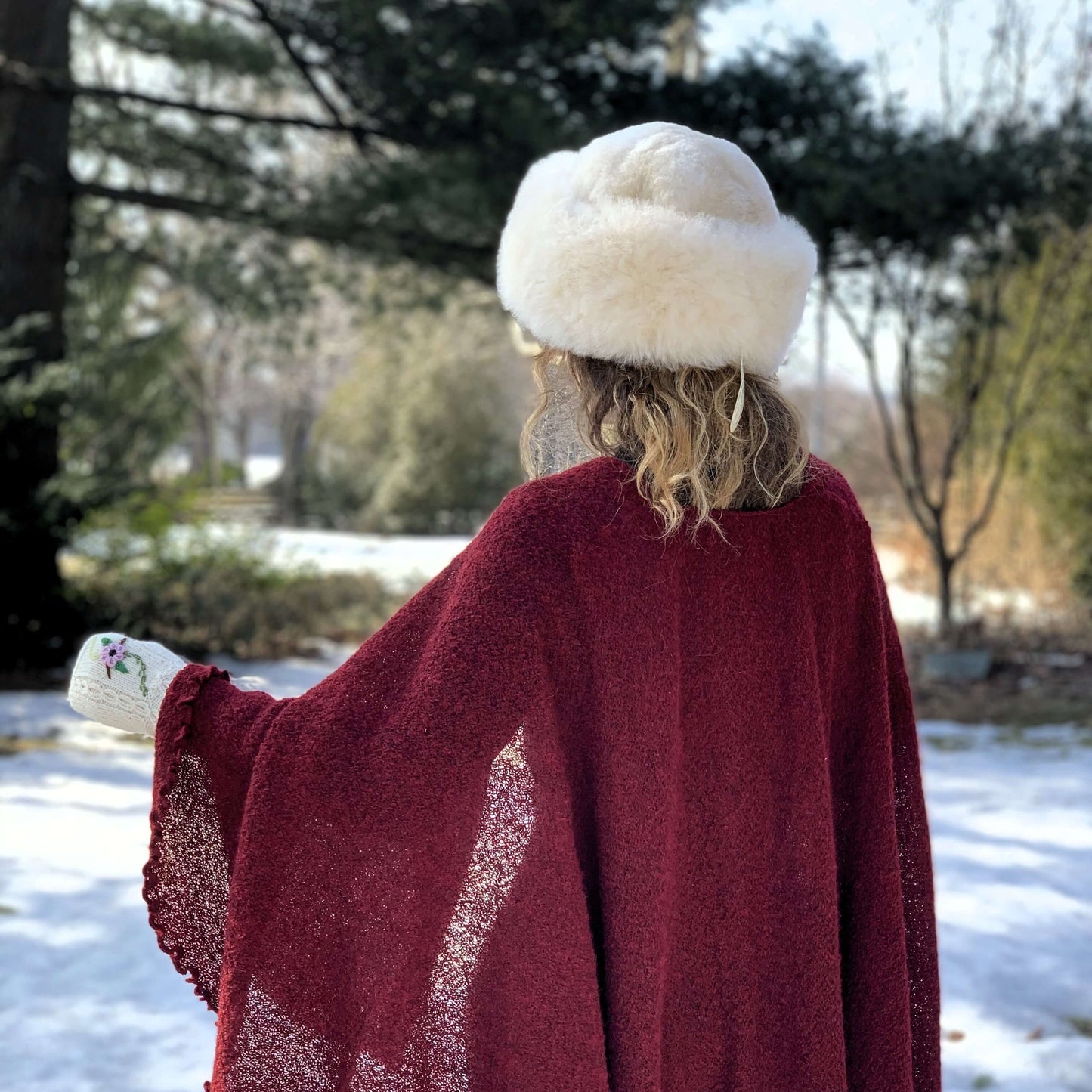 Back view of a woman wearing light beige Russian style alpaca fur hat and a burgundy cape with a winter background.