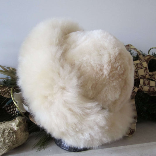 Back view of a light beige Russian style alpaca fur hat with greenery in the background.