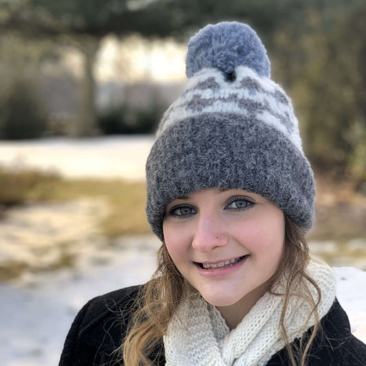 Woman wearing grey and ivory knit alpaca hat with large fluffy pompom with blurred natural background.