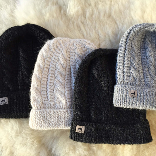 Four cable knit alpaca hats in black, ivory, charcoal, and silver grey on a fluffy white surface.