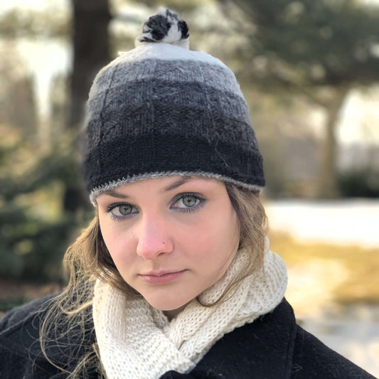 Woman wearing a patterned gray knit alpaca hat with a pom-pom and a white alpaca scarf outdoors.