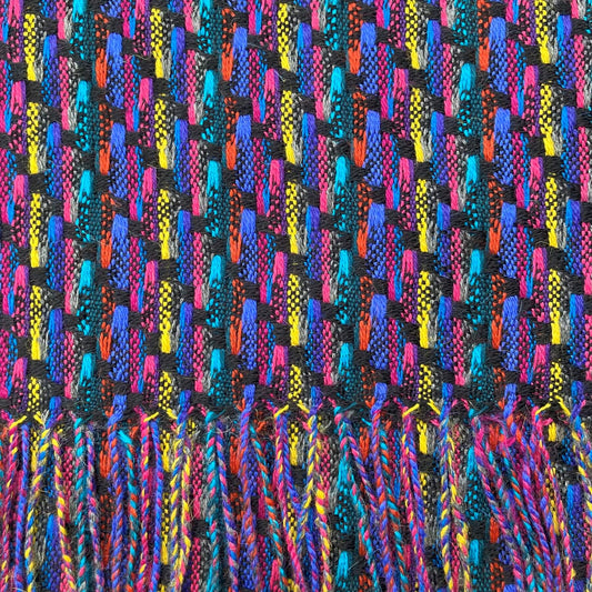 A close-up of a woven textile with a multicolor chevron pattern in bright blue, pink, yellow, orange, and teal on a black base, finished with a fringe tassel edge at the bottom.