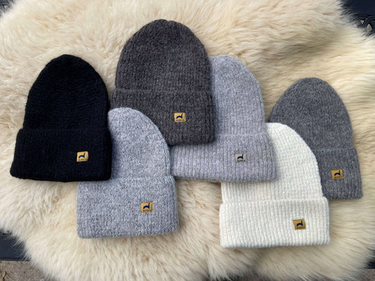 Six ribbed alpaca hats in black, ivory, and shades of grey with labels on a fluffy background.