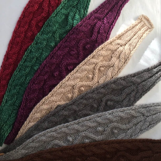 Seven cable knit alpaca headbands in burgundy, forest green, purple, beige, charcoal grey, silver grey, and brown.