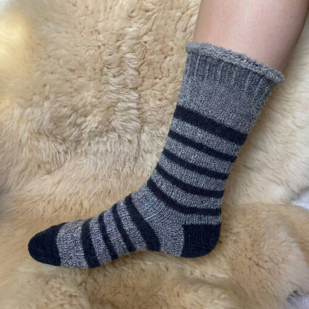 A single sock worn on a person's foot, resting on a soft beige alpaca fur surface. The sock is heather gray with black horizontal stripes and a black toe.