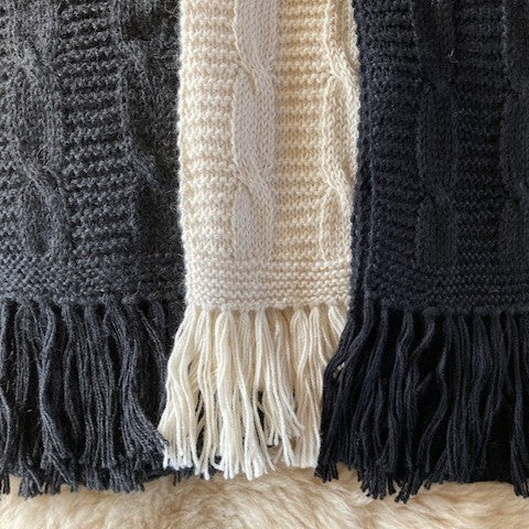 Close-up of a knitted cable-pattern scarves showing three colorways: dark charcoal grey, cream, and black, with fringed ends resting on a soft beige fur surface.