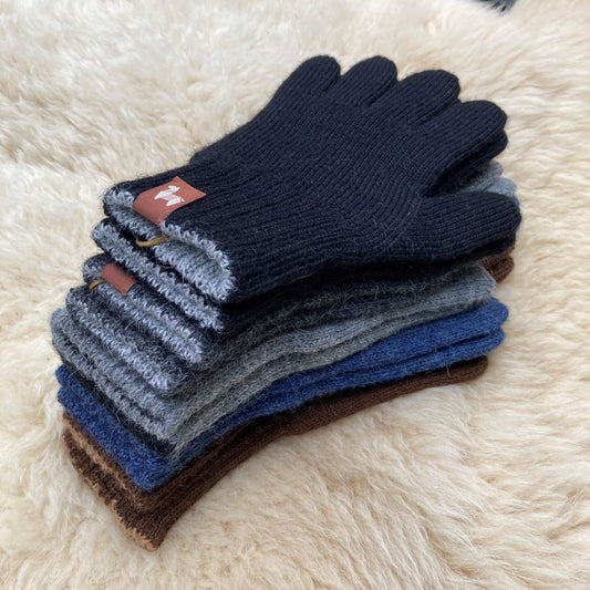A stack of ribbed knit gloves in multiple colors, with a black glove on top, placed on a soft cream fur surface. Small brown leather wrist tabs with white alpaca logos are visible.
