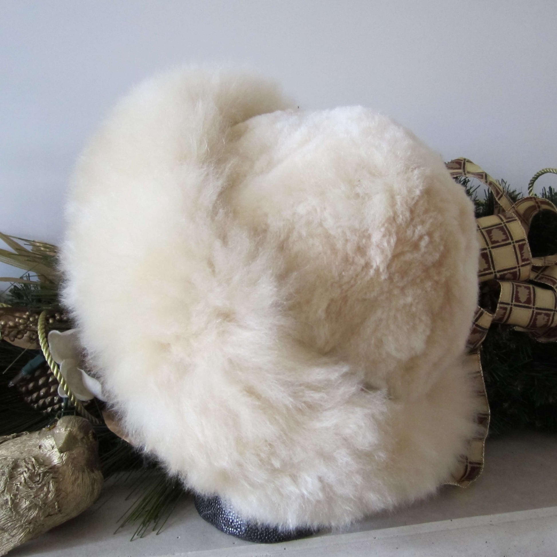 Back view of a light beige Russian style alpaca fur hat with greenery in the background.