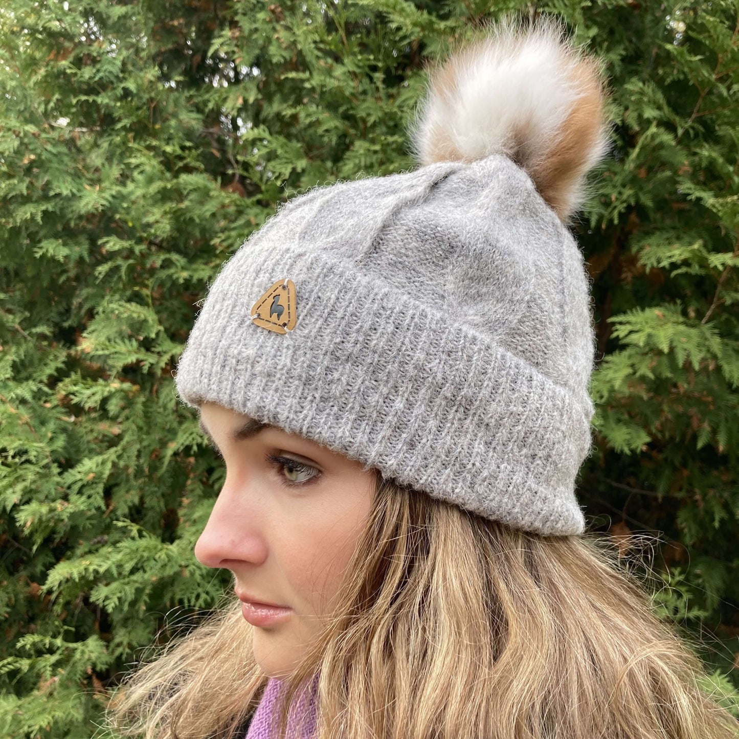 Woman wearing a silver grey cable knit alpaca hat with an alpaca fur pom-pom against a green foliage background