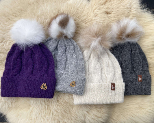 Four cable knit alpaca hats with alpaca fur pom-poms on a fluffy surface