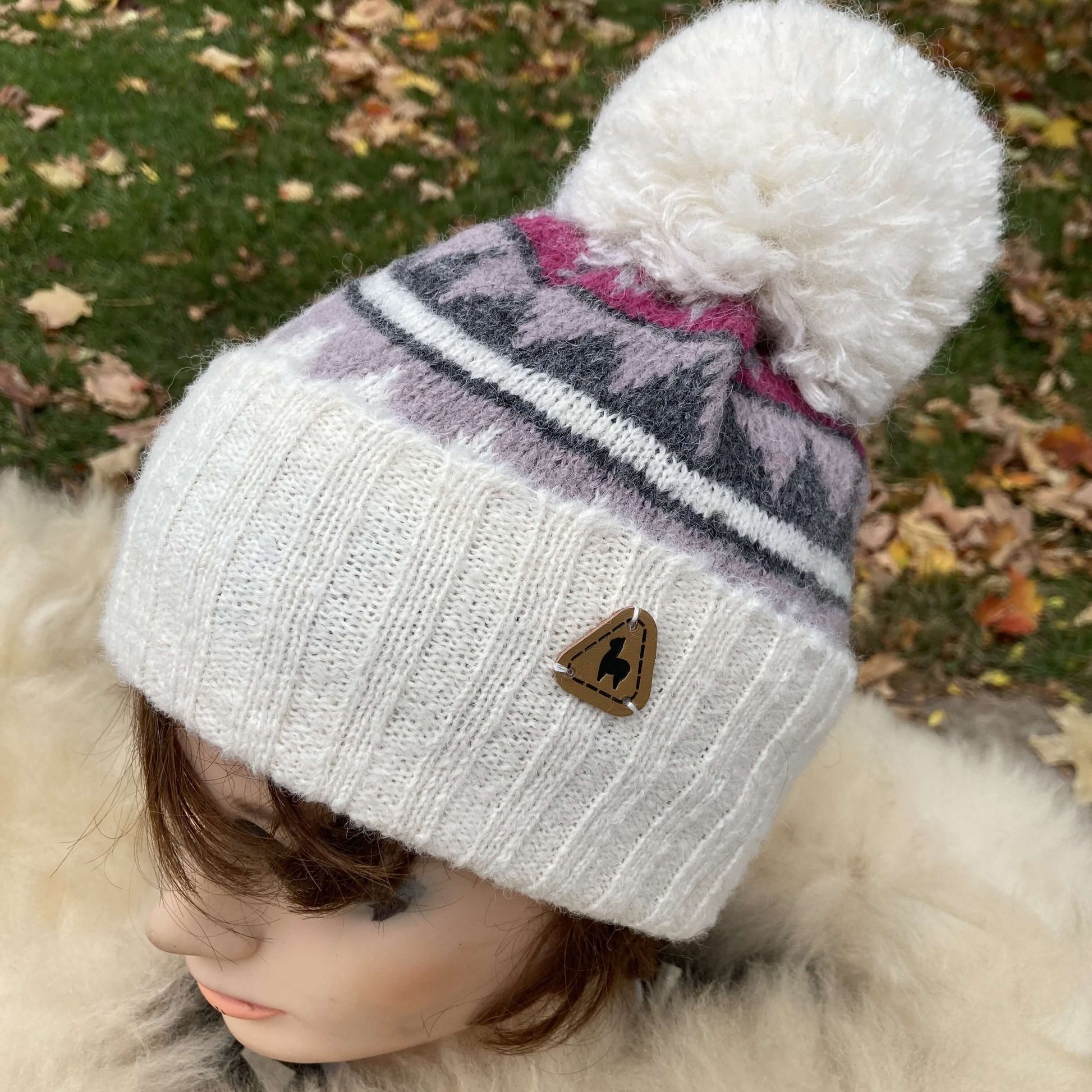 Mannequin head with ivory knit alpaca hat with grey, and magenta geometric patterns and a large fluffy pompom with autumn leaves in background.