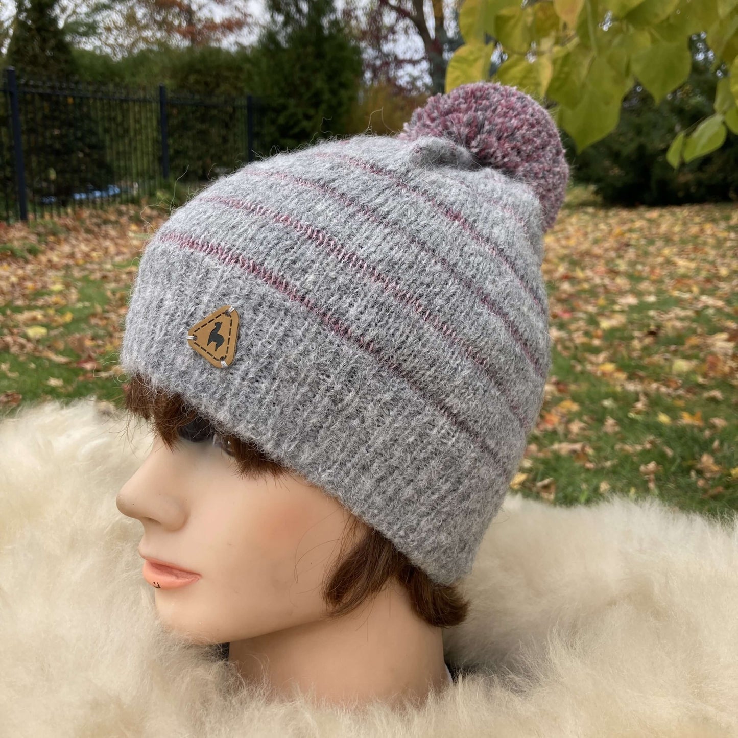 Mannequin head wearing light grey knit alpaca hat with large fluffy pompom and plum stripes with natural autumn background.