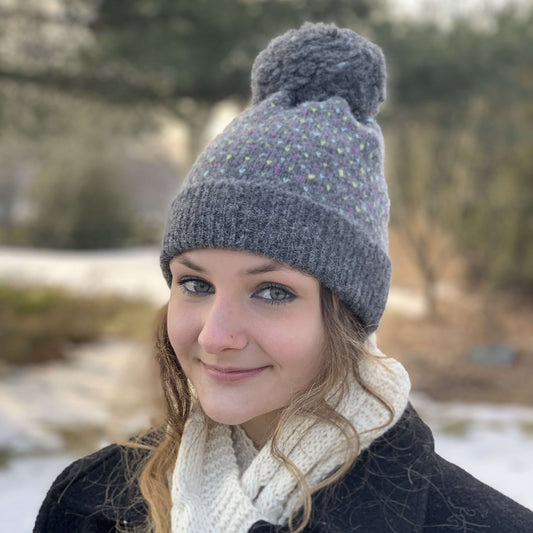Woman wearing grey knit alpaca hat with large fluffy pompom with a bllurred natural background.