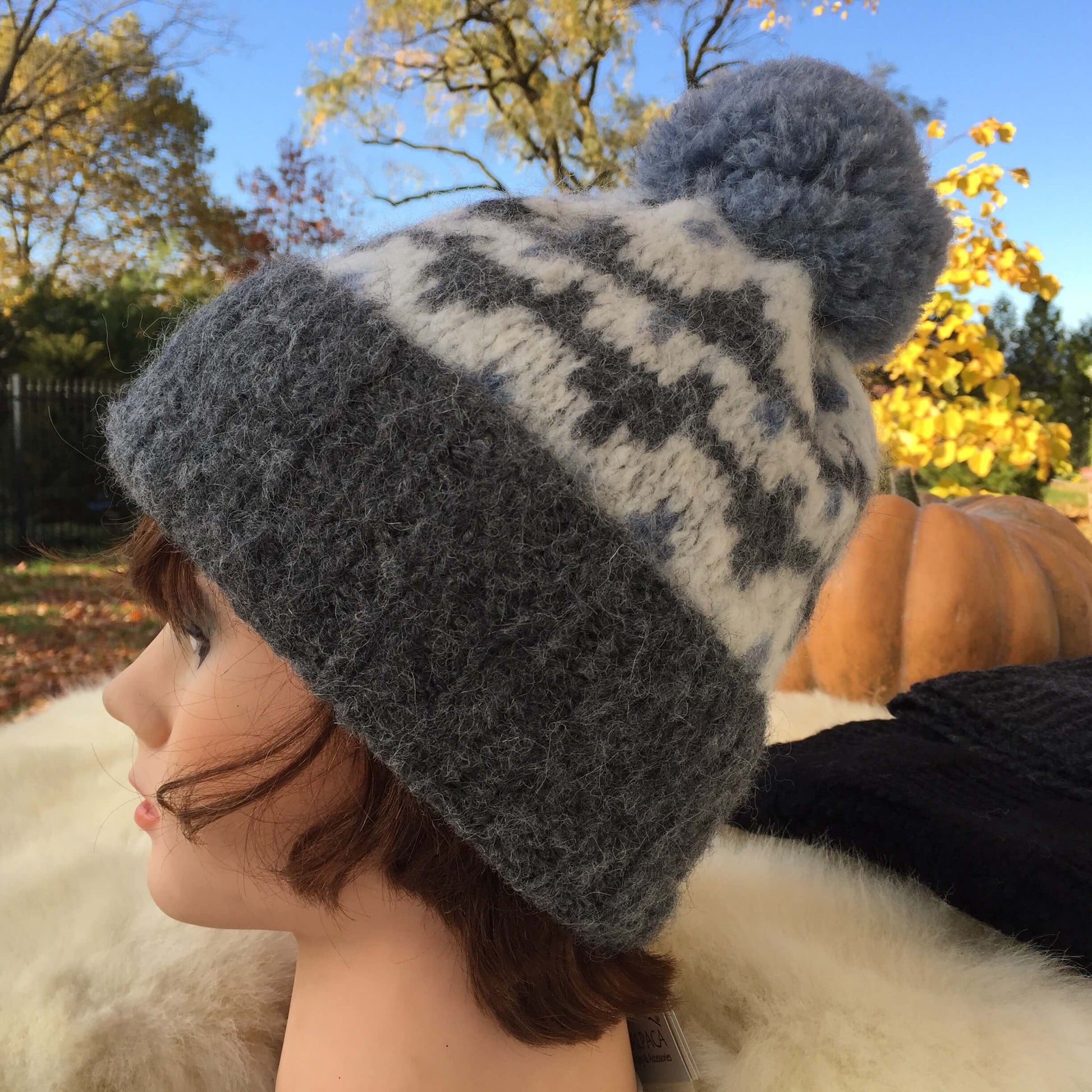 Mannequin head with charcoal grey knit alpaca hat with ivory and denim blue geometric patterns and a large fluffy pompom with autumn scene in background.