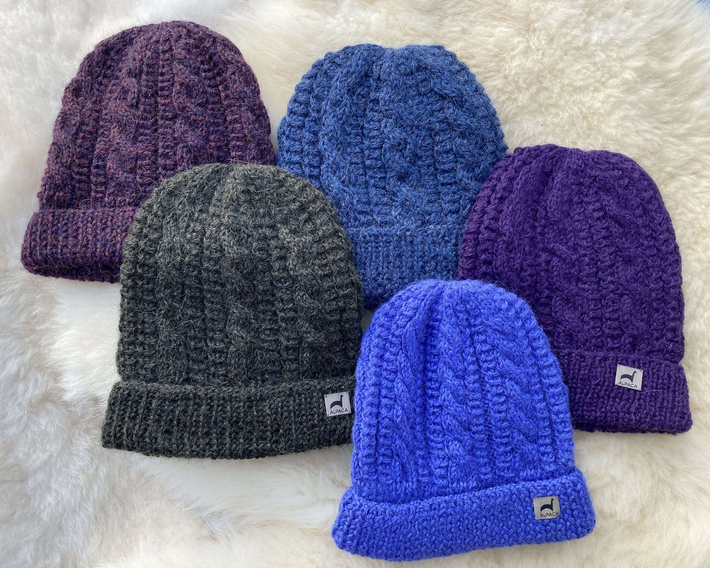 Five cable knit alpaca hats in purple, denim blue, and periwinkle on a white fluffy surface