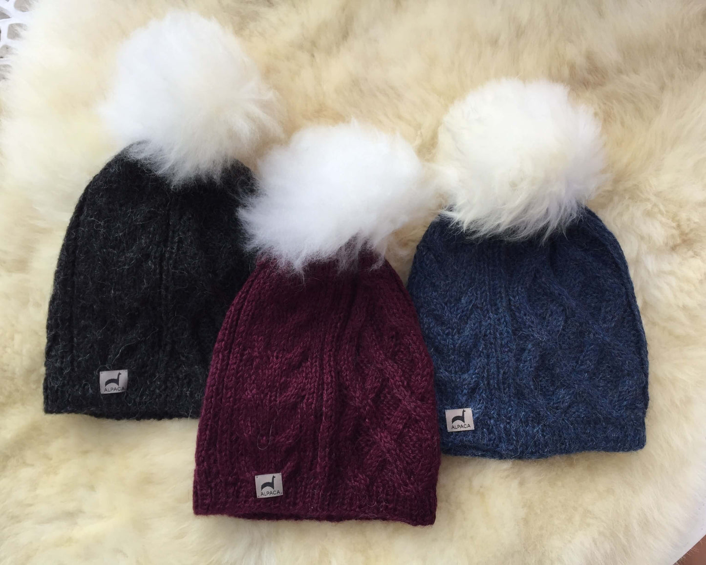 Three cable knit alpaca hats with alpaca fur pom-poms in charcoal, maroon, and blue on a fluffy white surface.
