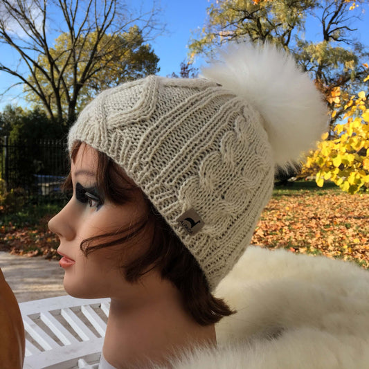 Cable Knit ivory beanie with a white alpaca fur pom-pom on a mannequin head against an outdoor background.