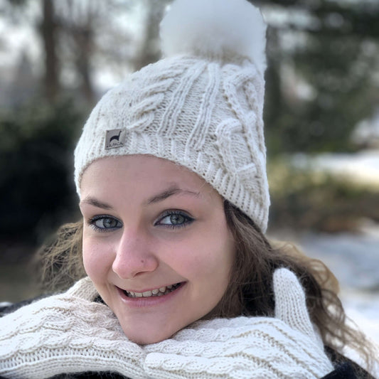 Woman wearing a white cable knit beanie with an alpaca fur pom-pom in a snowy outdoor setting