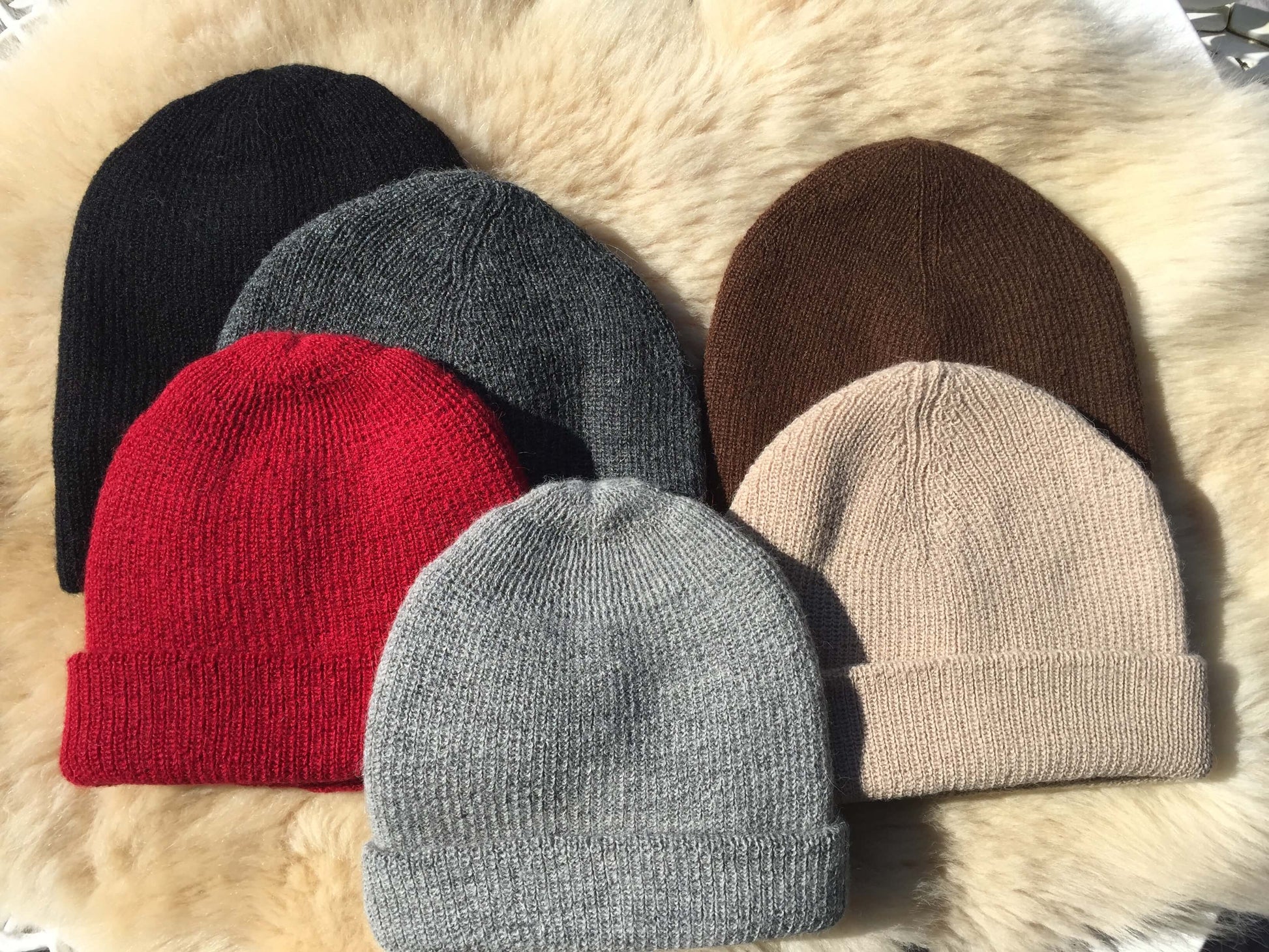 Six knit alpaca hats in black, gray, red, brown, and beige on a fur surface.