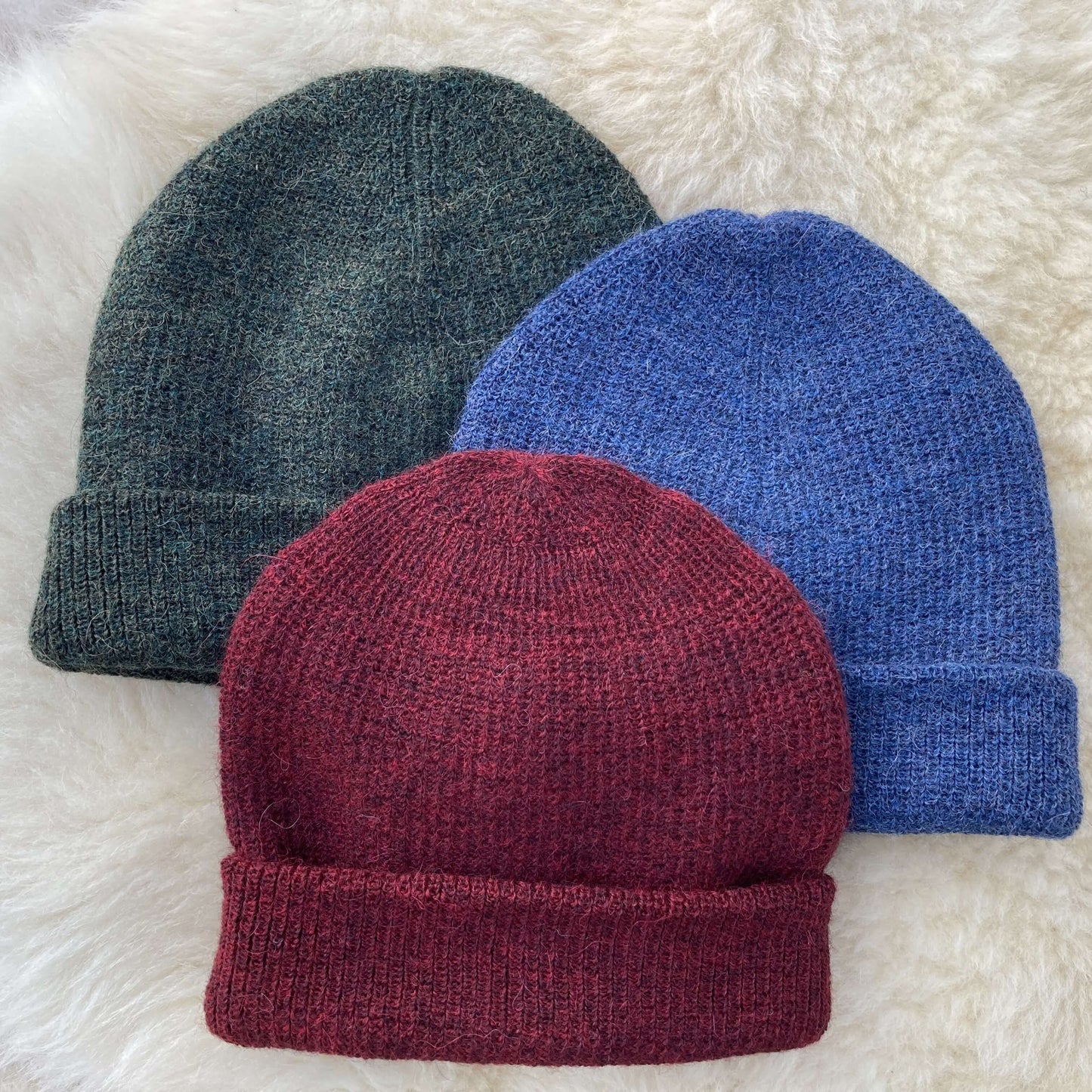 Three knit alpaca hats in burgundy, denim blue, and grey on a white fluffy surface.