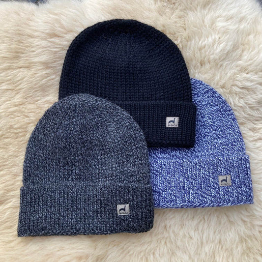 Three rib knit alpaca hats in black, gray, and blue on a fluffy white surface.