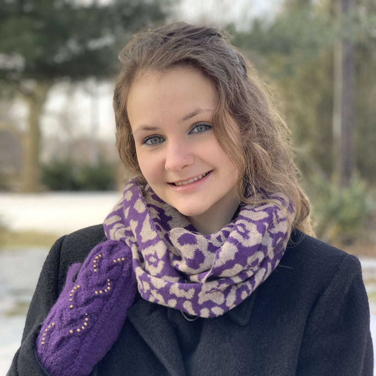 Young woman wearing a purple and taupe fabric scarf wrapped around her neck with a dark coat and purple mittens with a blurred nature background.