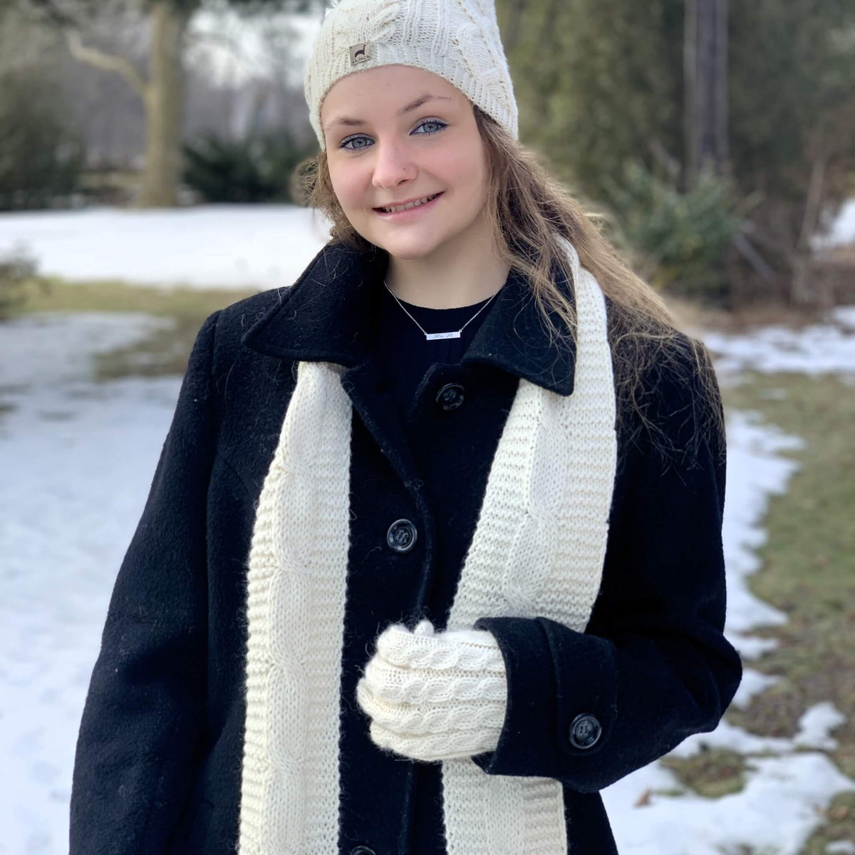 A young woman wearing a black wool coat, a thick cream cable-knit scarf, a matching cream knit beanie, and cream knit gloves, standing outdoors in a snowy setting.