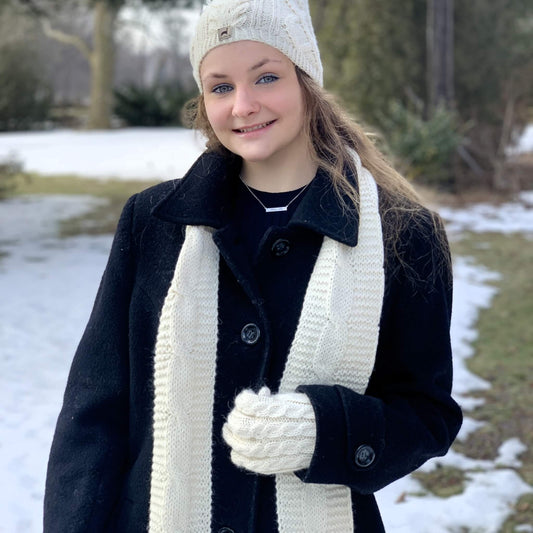 A young woman wearing a black wool coat, a thick cream cable-knit scarf, a matching cream knit beanie, and cream knit gloves, standing outdoors in a snowy setting.