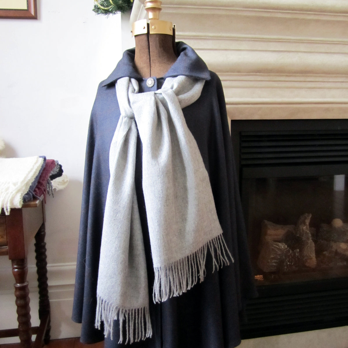 A light gray woven scarf with fringe ends draped over a dark navy blue cape displayed on a mannequin torso in an indoor room setting with a fireplace visible in the background.