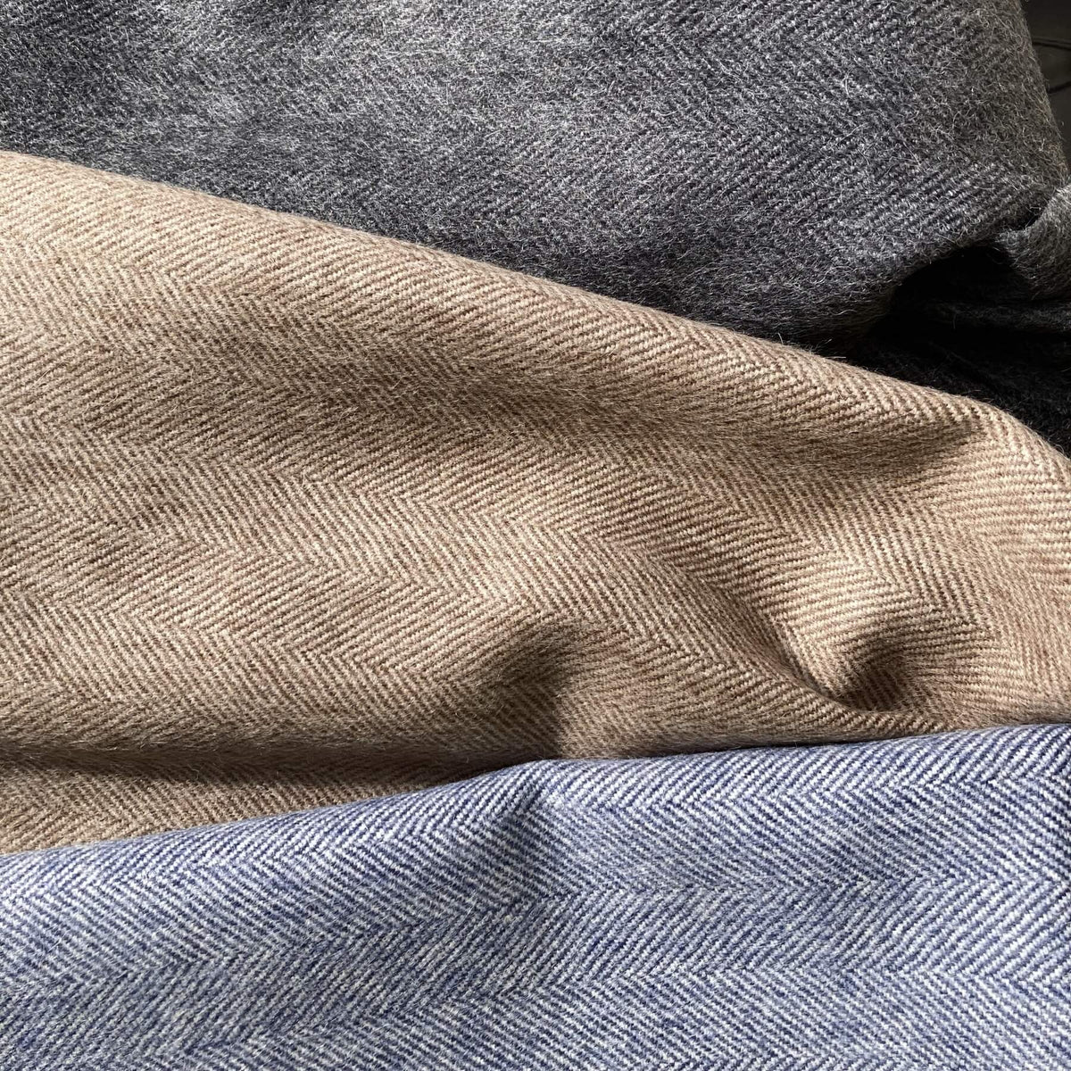 Three folded herringbone wool fabrics in gray, beige, and light blue, stacked diagonally to show their texture and color.