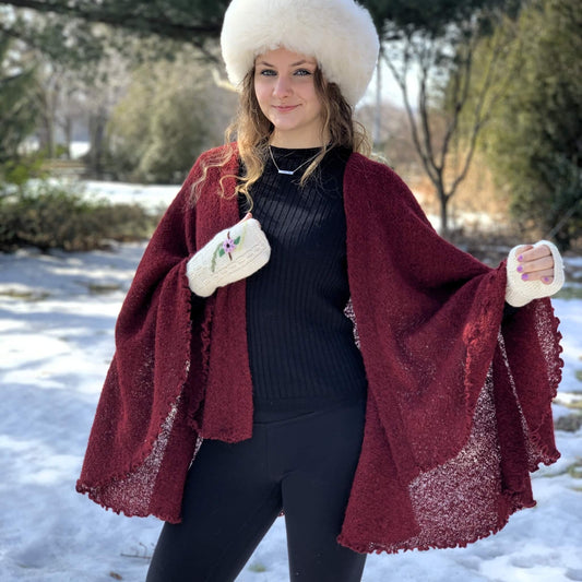 A woman wearing a burgundy open-front knit cape, a white fur hat, and white knit mittens, standing in a snowy outdoor setting.