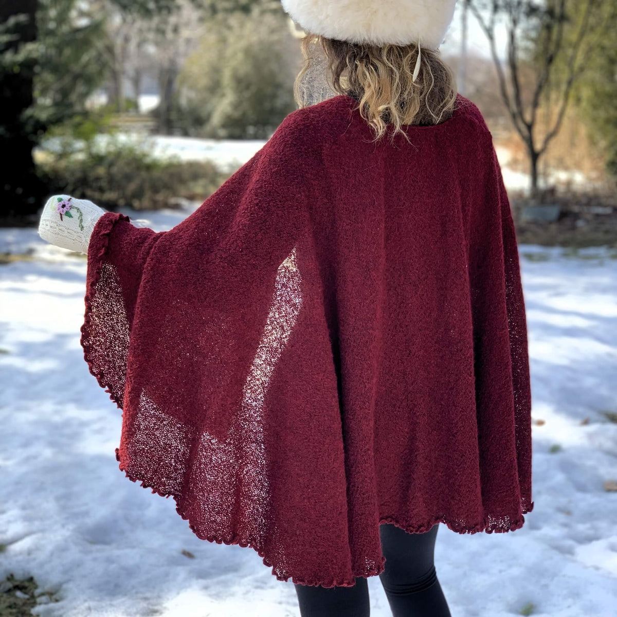 A person standing outdoors in the snow, shown from the back, wearing a loose burgundy knit cape with wide draped sleeves and scalloped edges. The person also wears a cream hat and white gloves.