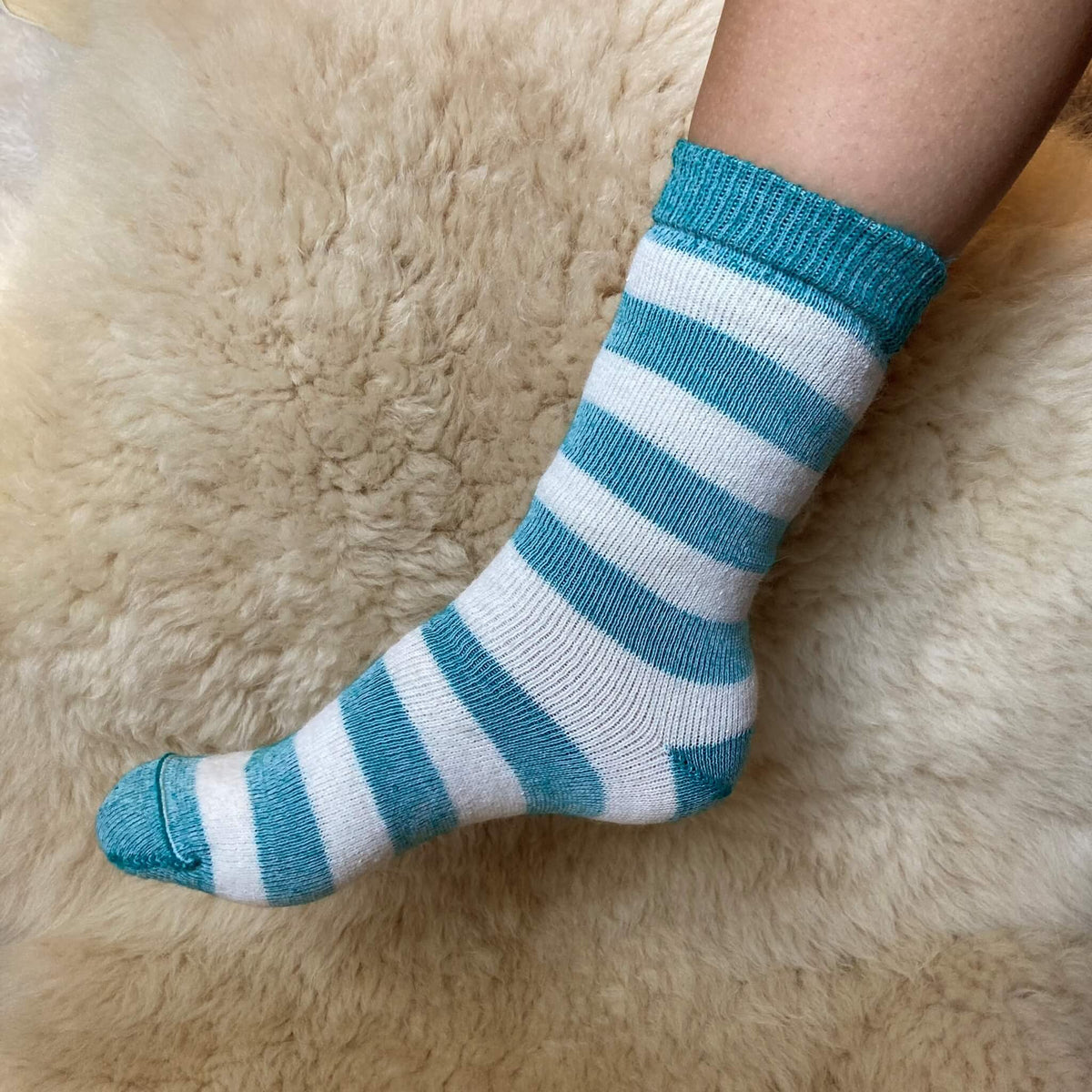 A person's foot wearing a knit crew sock with horizontal teal green and white stripes, resting on a soft beige alpaca fur surface.