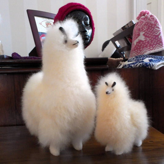 Two fluffy white alpaca-like stuffed animals standing on a dark wooden table in an indoor room. One is larger and the smaller one stands beside it. Alpaca hats are displayed in the background.