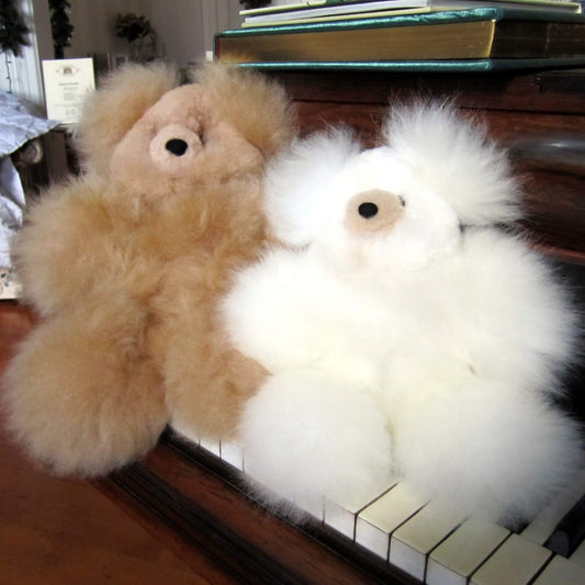 Two fluffy teddy bears made from alpaca fur, one tan and one white, sitting on a piano keyboard in an indoor setting.