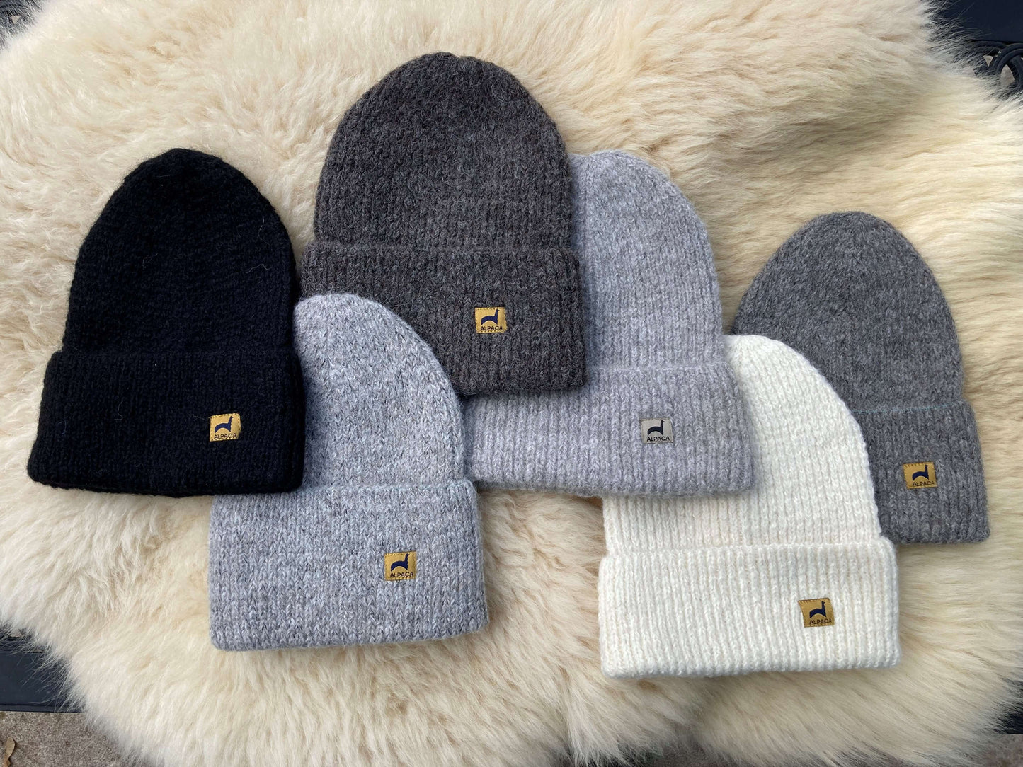 Six ribbed alpaca hats in black, ivory, and shades of grey with labels on a fluffy background.