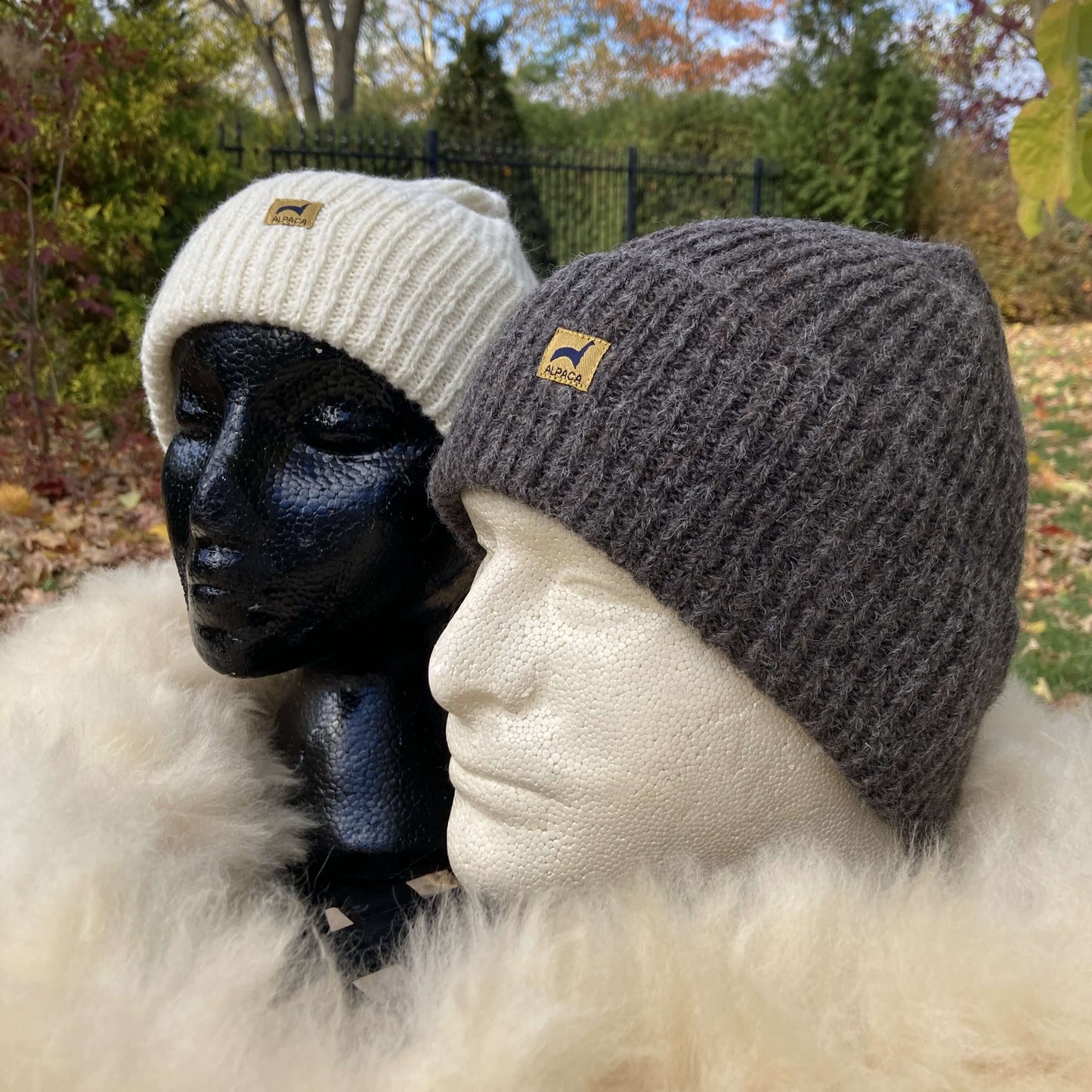 A female mannequin head wearing an ivory ribbed alpaca hat and a male mannequin head wearing a grey ribbed alpaca hat surrounded by a fluffy scarf and a natural autumn background.