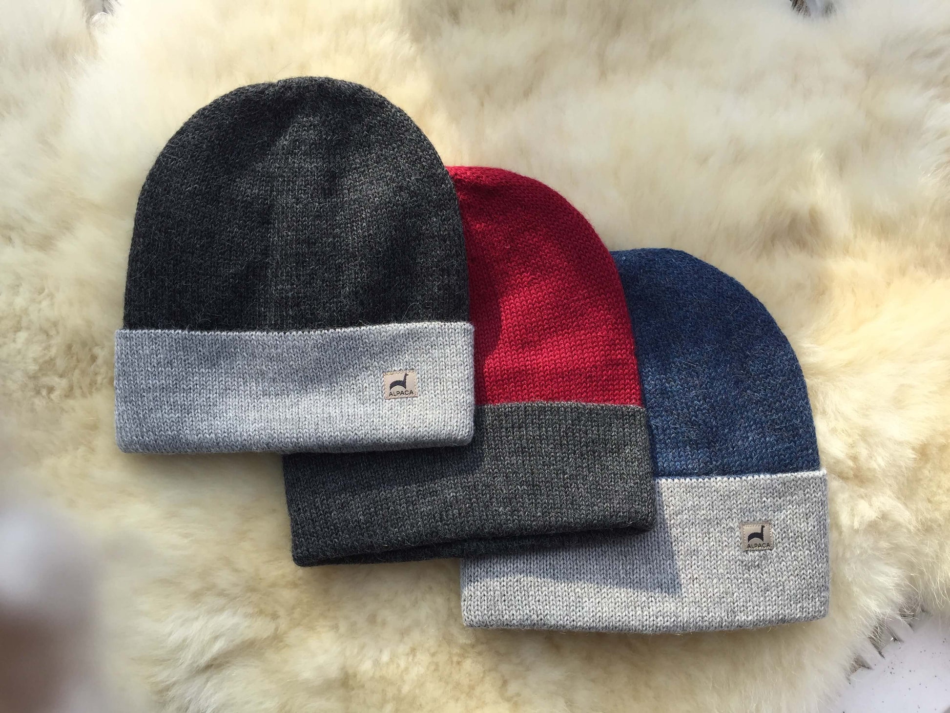 Three knit alpaca hats in charcoal grey, burgundy, and denim blue on a fluffy beige surface.