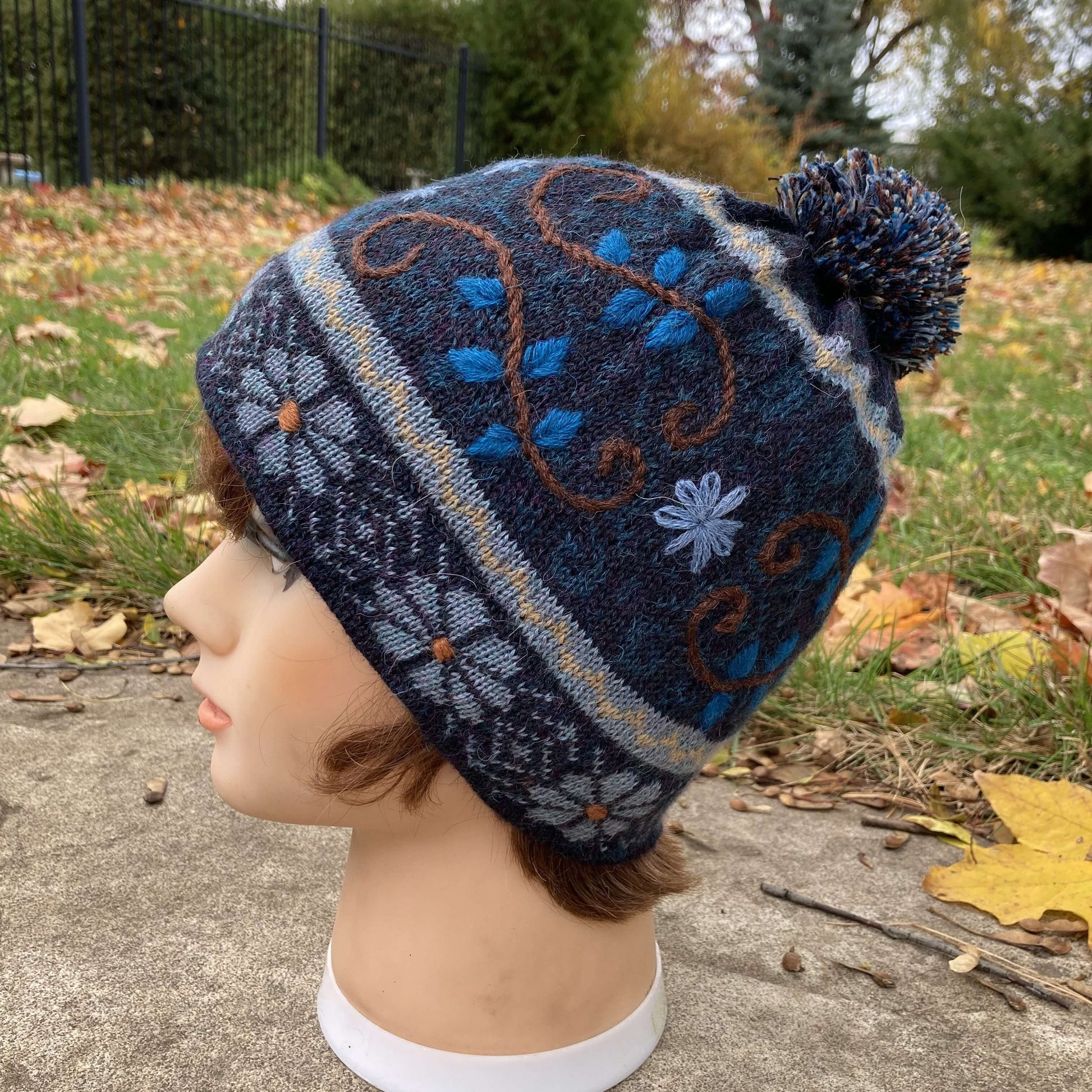 Female mannequin head wearing a dark  blue, light blue, and brown embroidered knit alpaca hat with a pompom with an autumn scene in the background.