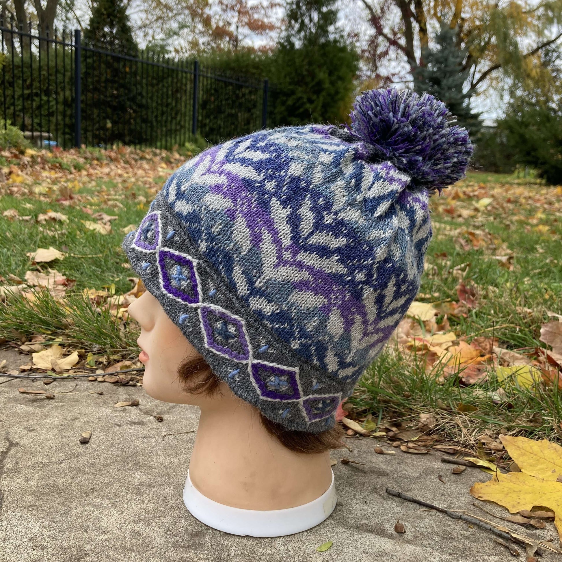 Female mannequin head wearing a light grey, purple, and blue embroidered knit alpaca hat with a pompom with an autumn scene in the background.