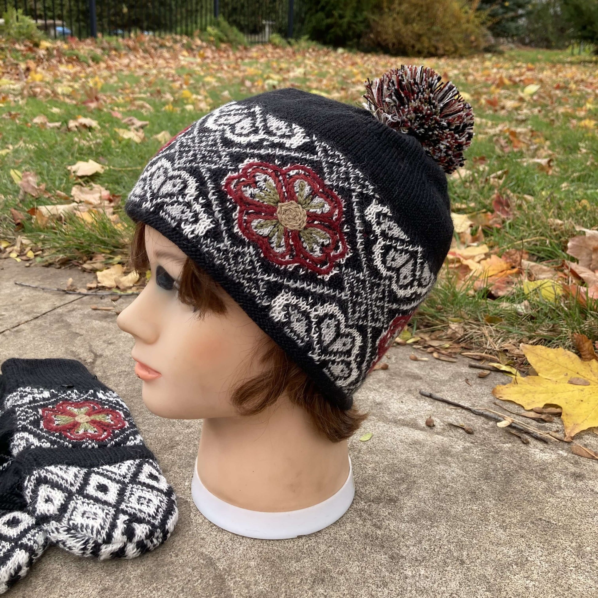 Female mannequin head wearing a black, burgundy, and ivory embroidered knit alpaca hat with a pompom with an autumn scene in the background.