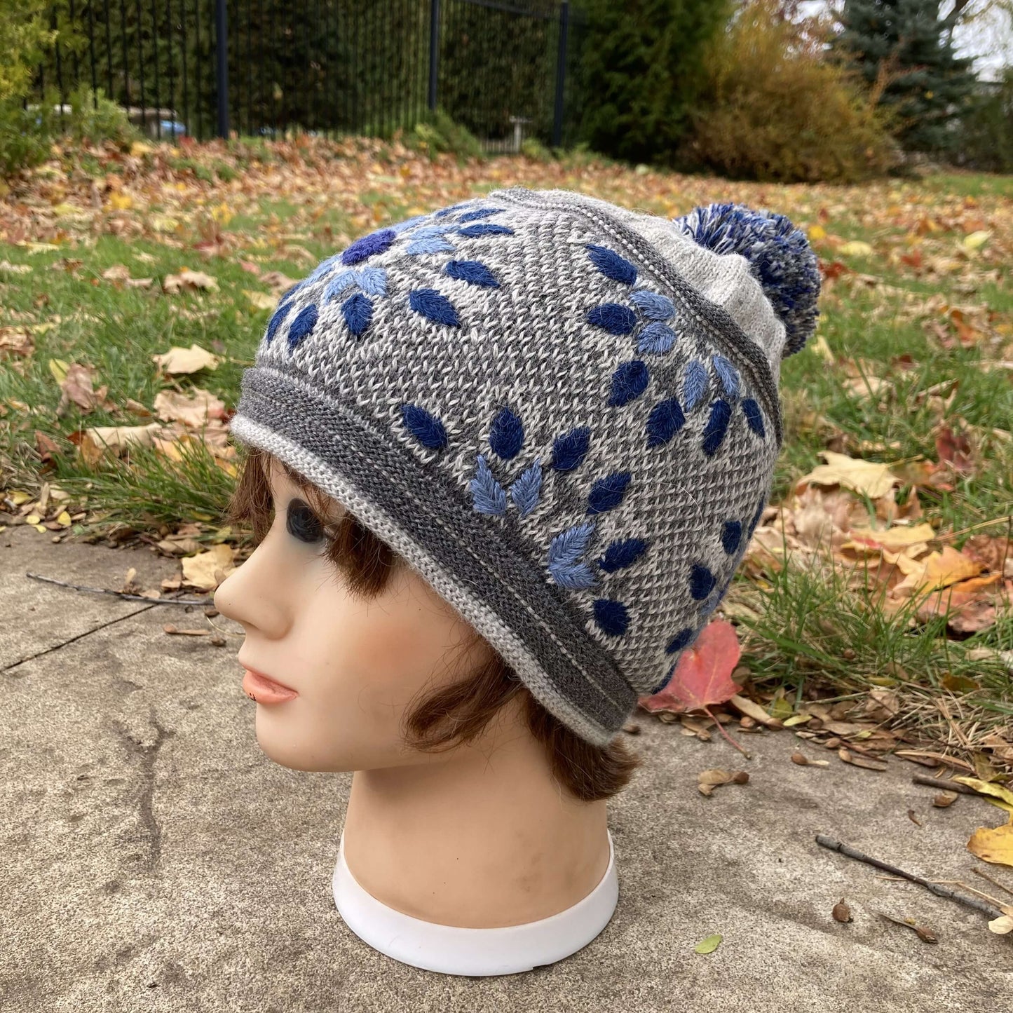 Female mannequin head wearing a light grey, navy, and denim blue embroidered knit alpaca hat with a pompom with an autumn scene in the background.