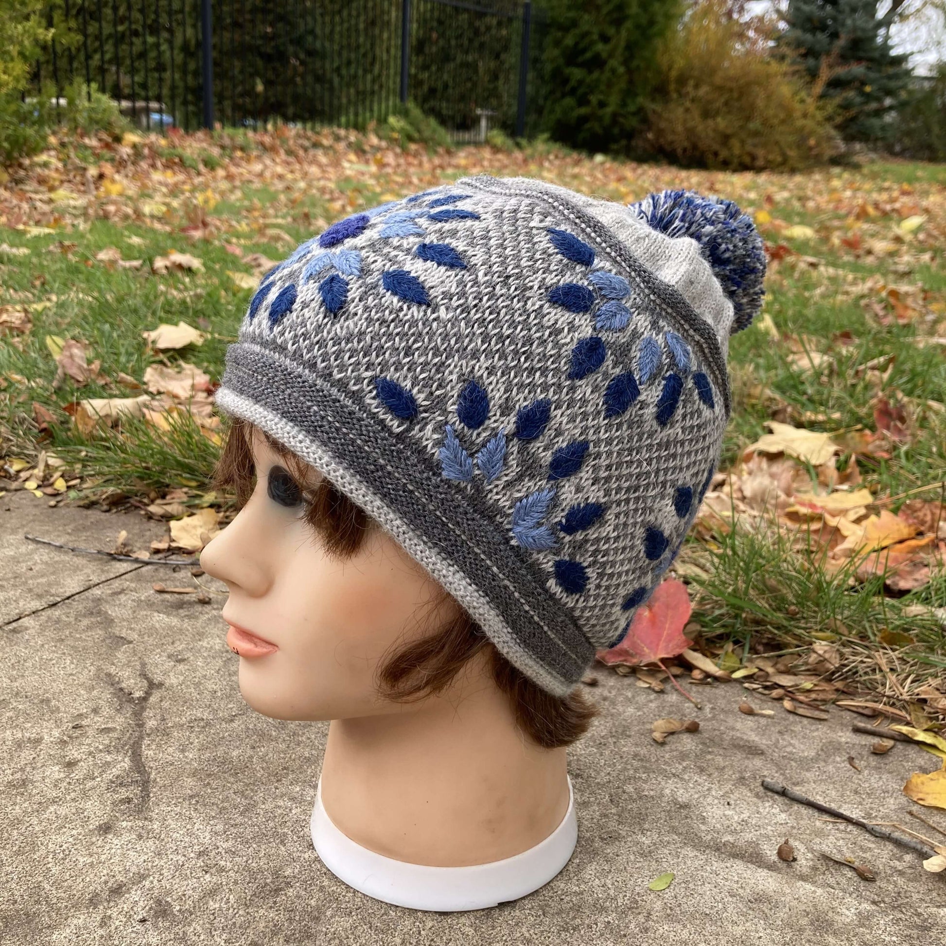 Female mannequin head wearing a light grey, navy, and denim blue embroidered knit alpaca hat with a pompom with an autumn scene in the background.