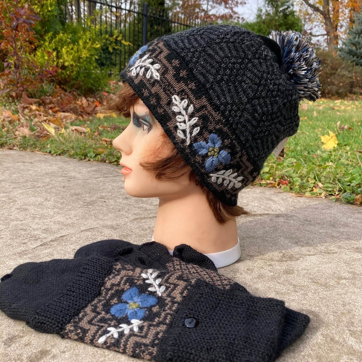 Female mannequin head wearing a black, taupe, and ivory embroidered knit alpaca hat with a pompom with an autumn scene in the background.