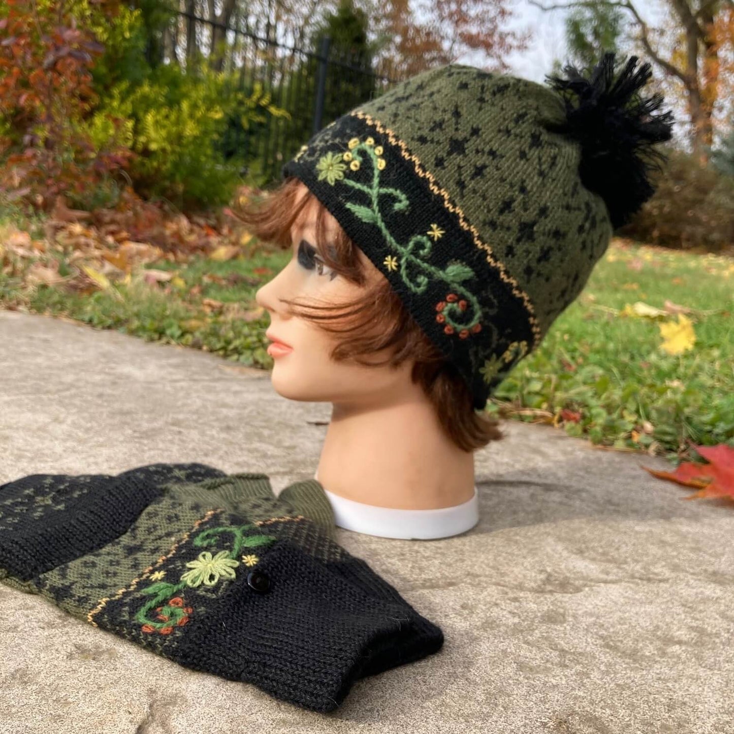 Female mannequin head wearing a black and green embroidered knit alpaca hat with black pompom with an autumn scene in the background.