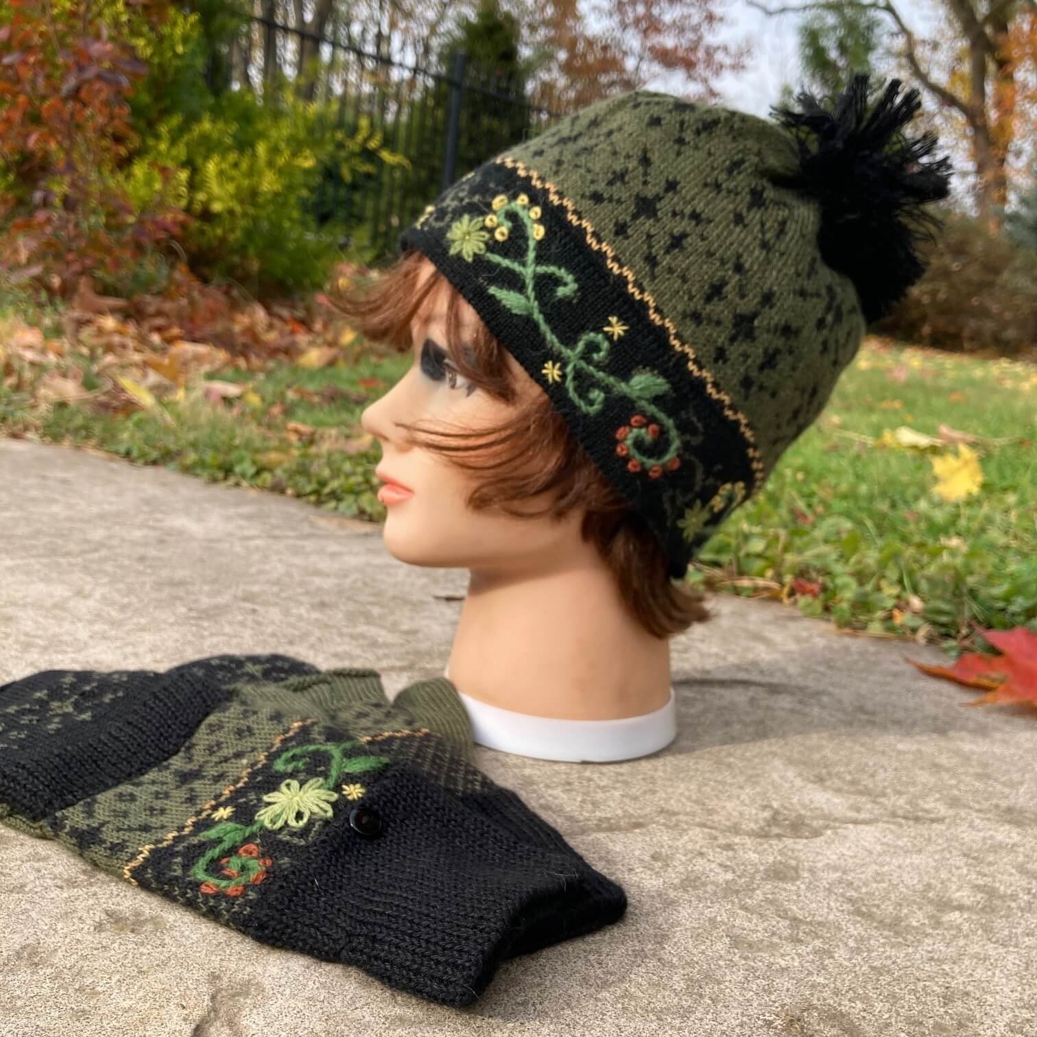Female mannequin head wearing a black and green embroidered knit alpaca hat with black pompom with an autumn scene in the background.