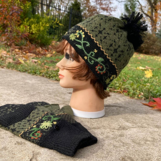 Female mannequin head wearing a black and green embroidered knit alpaca hat with black pompom with an autumn scene in the background.