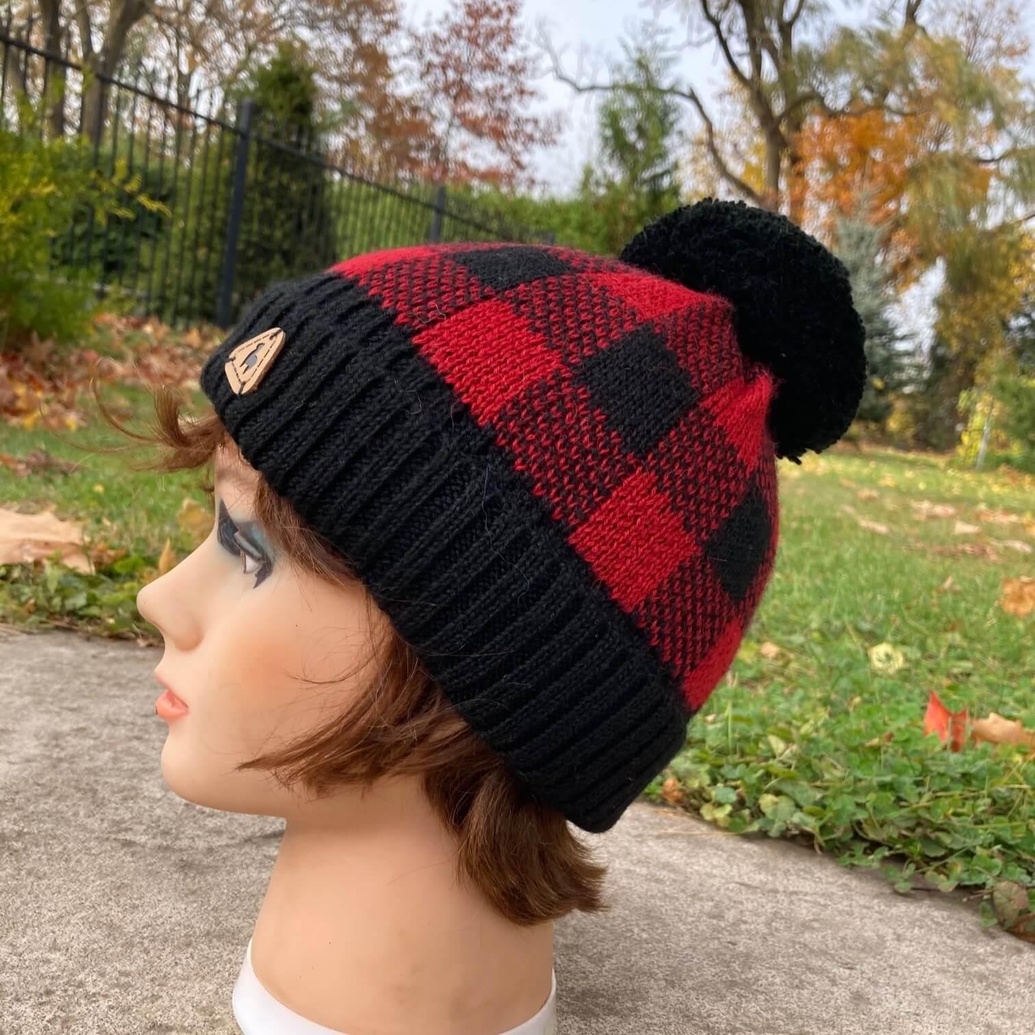 Female man nequin head wearing a black and red plaid knit alpaca hat with a black pompom with an autumn scene in the background.