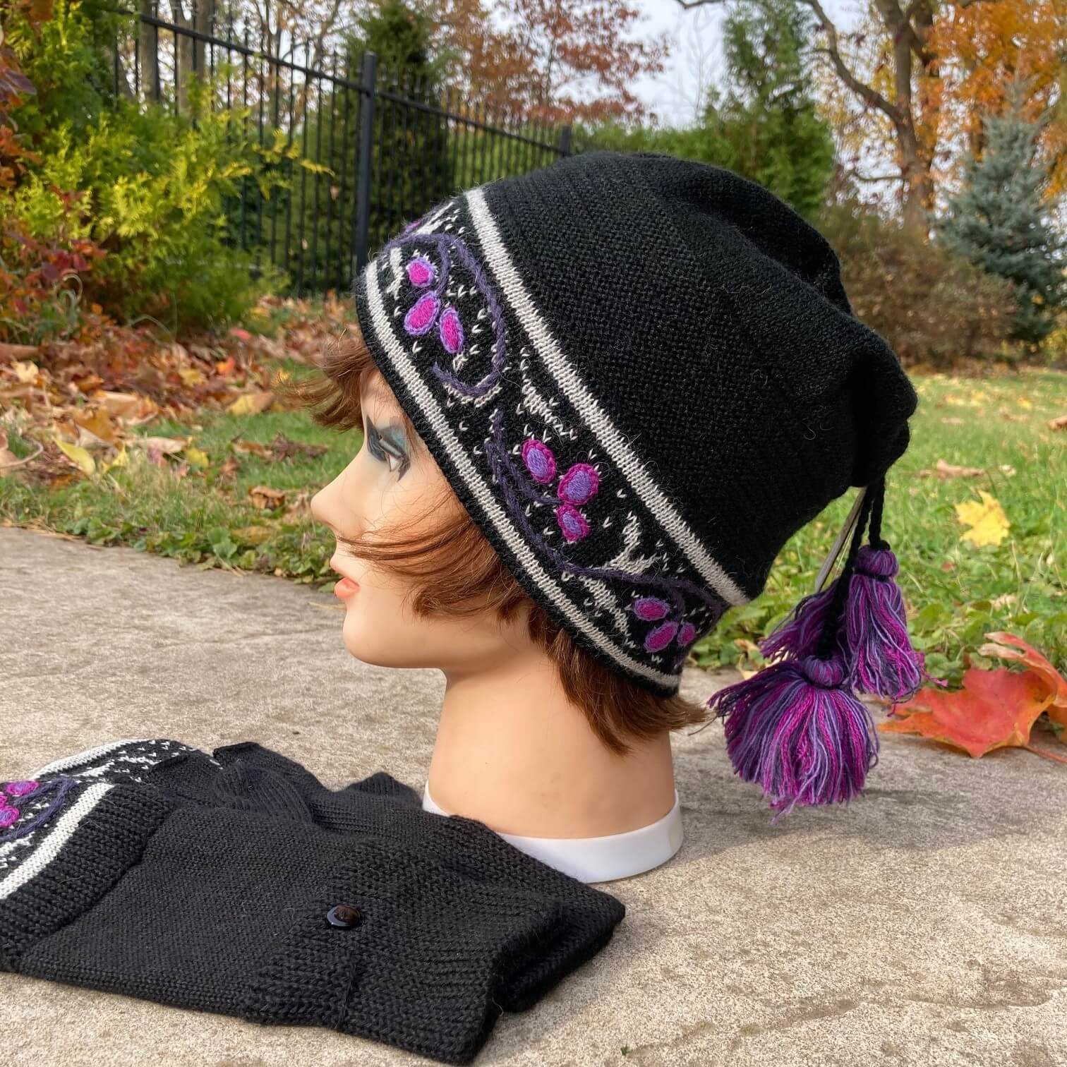 Female mannequin head wearing a black, purple, and ivory embroidered knit alpaca hat with a tassle with an autumn scene in the background.