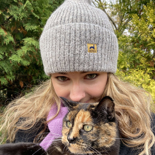 Woman wearing light grey ribbed alpaca hat with label holding a torti cat with evergreens in background.